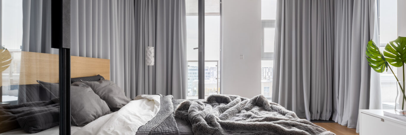Gallery Bedroom interior with gray curtains, a light-colored bed, and a plant by the window.