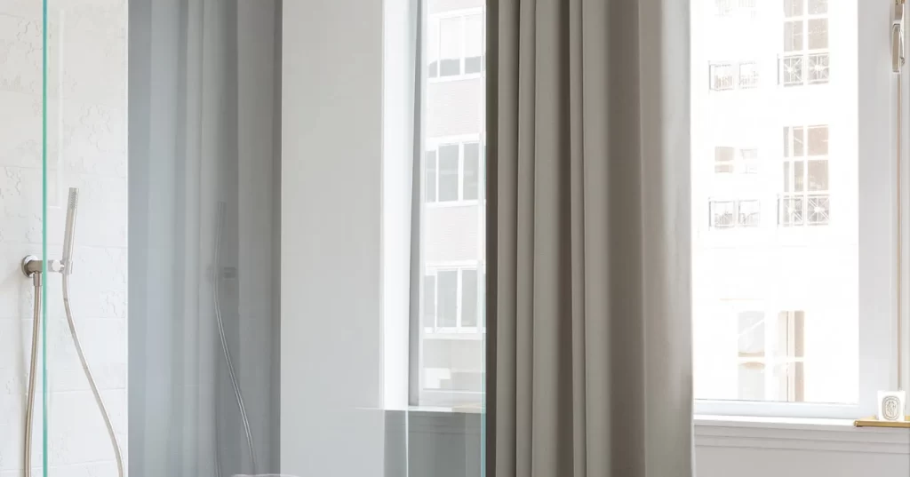 gallery A modern, minimalist shower stall. The stall is enclosed by clear glass panels, and light gray-beige curtains hang from the shower's frame.