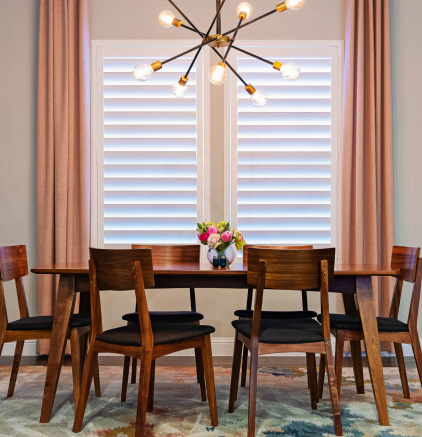 Gallery Wooden dining table with six chairs, flower vase centerpiece, modern chandelier, window with blinds and curtains.