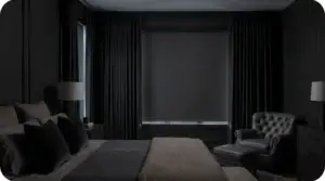 A calm, dark bedroom with a blackout roller shade and drapery keeping the room shaded against bright morning light outside