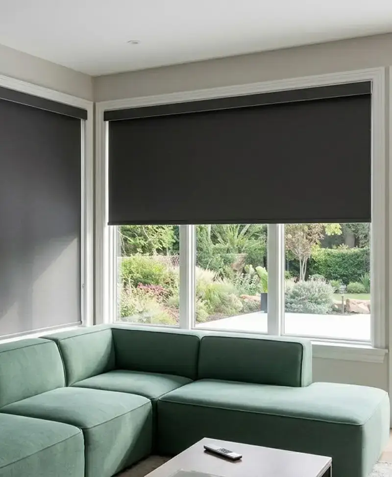 White roller shade lowered over a black-framed window beside a white lounge chair
